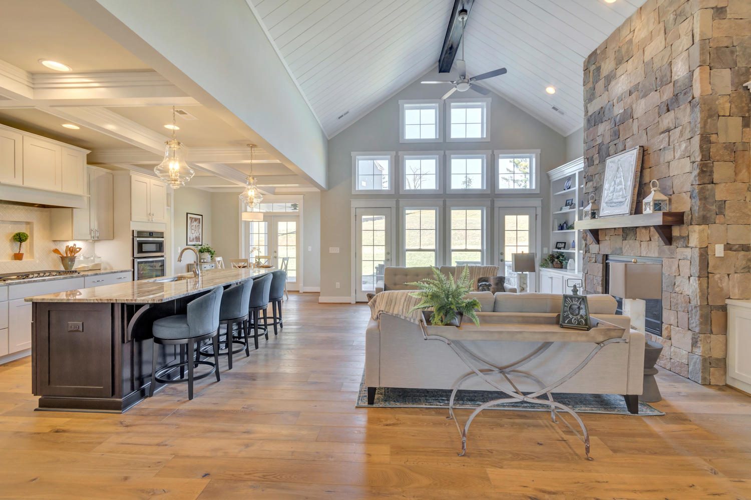 An open concept kitchen and great room with floor to ceiling windows. A comfortable living space centered around a stone fireplace is on the right, with a vaulted ceiling and ceiling fan above it. The kitchen is to the left with an island with upholstered high chairs.